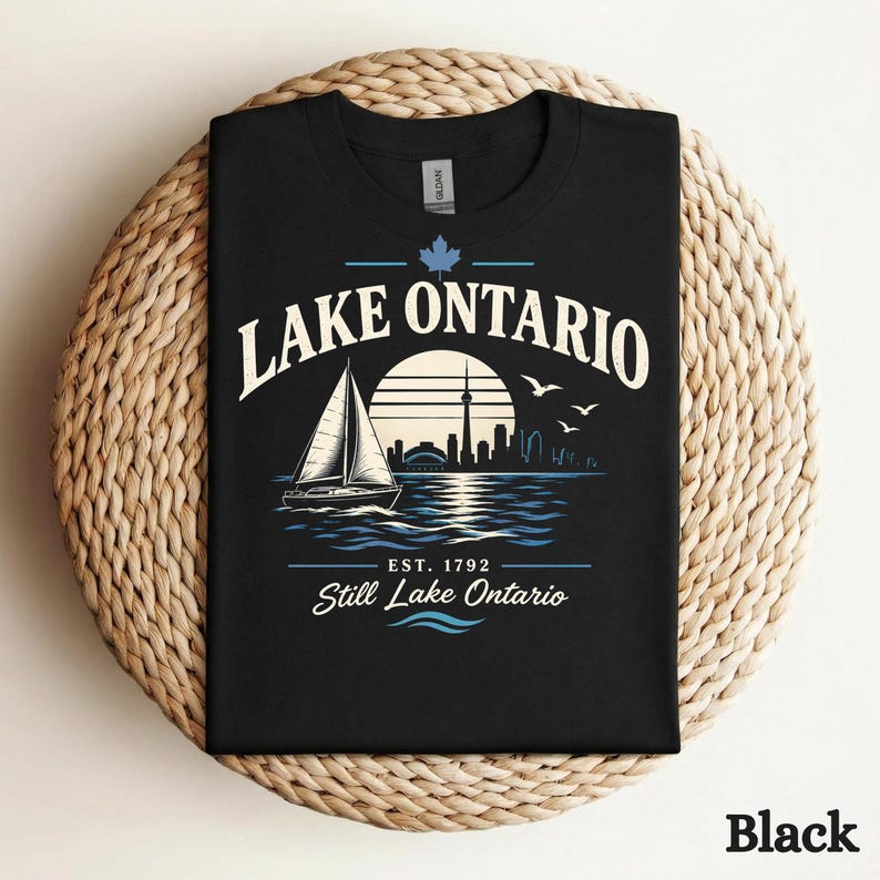 May include: A black t-shirt featuring a graphic of a sailboat on water with a city skyline silhouette, a sun, and birds. The text reads LAKE ONTARIO, EST. 1792, and Still Lake Ontario, with a small maple leaf icon above the text.