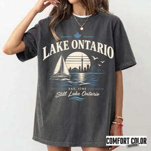 May include: A charcoal gray short-sleeve t-shirt featuring a graphic print with the text LAKE ONTARIO, a sailboat, a sun, a city skyline, and birds. Below the graphic, the text reads EST. 1792 and Still Lake Ontario in white and blue ink.
