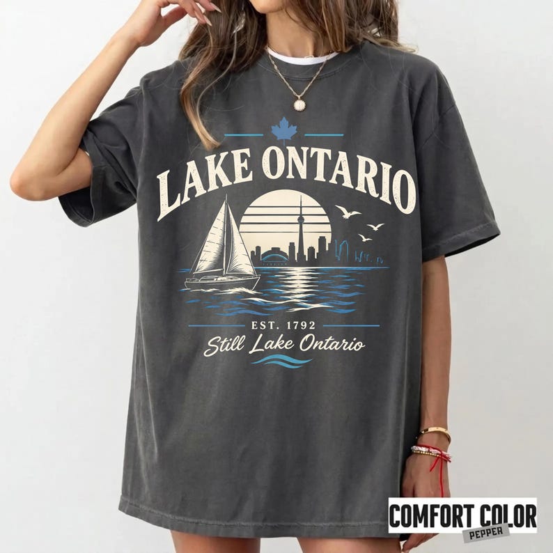 May include: A charcoal gray short-sleeve t-shirt featuring a graphic print with the text LAKE ONTARIO, a sailboat, a sun, a city skyline, and birds. Below the graphic, the text reads EST. 1792 and Still Lake Ontario in white and blue ink.