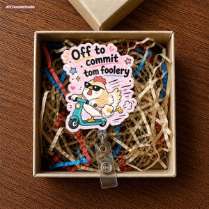 Può includere: Un portabadge con un pollo a cartoni animati che indossa occhiali da sole su un monopattino turchese. Lo sfondo rosa include il testo Off to commit tom foolery, piccole stelle e cuori. L'oggetto è contenuto in una scatola regalo di cartone.