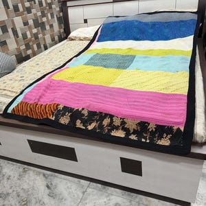 May include: A rectangular quilt featuring a patchwork design of horizontal fabric panels in blue, white, lime green, light blue, yellow, and pink. The quilt is finished with a black border and a bottom section featuring a black floral pattern.