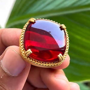 May include: A gold-toned ring featuring a large, square-cut, deep red translucent gemstone with rounded corners. The setting has a textured, beaded border and four prongs securing the stone. The metal has a matte, brushed finish throughout.
