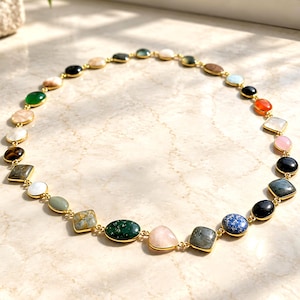 May include: A gold-tone chain necklace featuring a series of multi-colored, polished gemstones in various shapes, including round, oval, and square cuts. The stones display a diverse palette of colors such as green, blue, pink, black, and white.