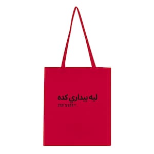 Op de afbeelding: Een felrode tote bag met twee lange schouderbanden. De voorzijde van de tas is voorzien van zwarte tekst in Arabisch schrift en de Engelse tekst "SNO NADRI" in een vet, schreefloos lettertype, gecentreerd op het onderste gedeelte van de tas.