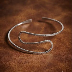 Könnte beinhalten: Ein silberfarbener Metall-Armreif mit einem offenen, fließenden, wellenartigen Design. Die Oberfläche des Metalls weist eine texturierte, gehämmerte Optik auf, die das Licht einfängt und ein dezentes Muster auf dem geschwungenen Band erzeugt.