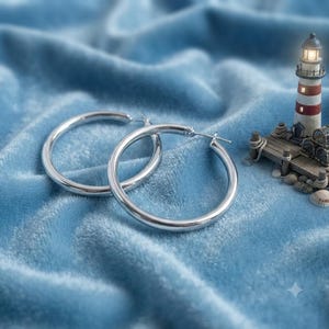 Könnte beinhalten: Ein Paar polierte silberne Creolen mit einem glatten, abgerundeten Profil und einem Klappverschluss. Die Ohrringe sind kreisförmig und verfügen über eine reflektierende metallische Oberfläche, die das Licht einfängt und schlicht wirkt.