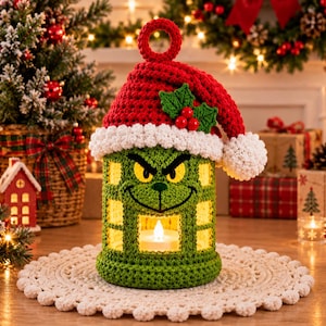 May include: A cylindrical green crochet lantern featuring a grumpy face with yellow eyes and a black stitched smile. The lantern is topped with a red and white crochet Santa hat adorned with green holly leaves and red berries, with a light inside.