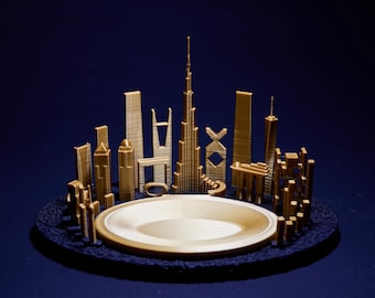 Op de afbeelding: Een goudkleurig 3D-geprint miniatuur skyline-model met diverse wolkenkrabbers in een halve cirkel rondom een centraal cirkelvormig platform. De structuren omvatten een hoge centrale toren en verschillende geometrische gebouwen op een donkere basis.