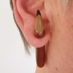 May include: A brown and black pencil-shaped ear gauge. The gauge is made of wood and has a black tip. The gauge is worn in a person's ear.