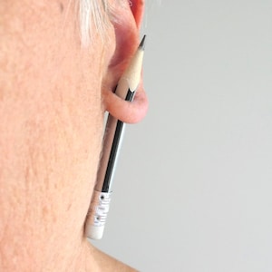 Könnte beinhalten: Ein silberfarbener Bleistift-Ohrring mit einer realistischen Graphitspitze und einem weißen Radiergummi am Ende. Der Bleistift ist so gestaltet, dass er aussieht, als würde er durch das Ohrläppchen verlaufen, mit einer glänzenden Metalloberfläche.