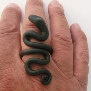 May include: A black snake-shaped ring made of polymer clay. The ring is worn on the middle finger of a hand.