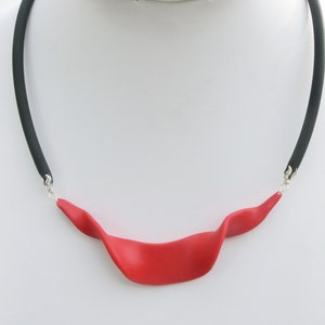 Könnte beinhalten: Eine Halskette aus roter Polymer-Knete mit einem schwarzen Gummiband. Die Halskette hat ein einziges, langes, gebogenes Stück aus roter Knete.