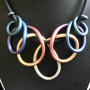 Op de afbeelding: Een veelkleurige metalen ketting met een reeks in elkaar grijpende ringen. De ringen zijn in tinten blauw, paars en goud.
