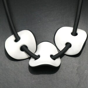 May include: A modern necklace featuring three white, rounded pendants with black edges and circular holes, strung on a black cord. The necklace has a minimalist design and is set against a dark gray background.