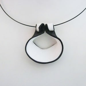 Peut inclure: Un collier en céramique noir et blanc avec un pendentif unique et abstrait. Le pendentif a la forme d'une boucle avec un trou au centre. Le collier est fait d'un cordon noir fin.