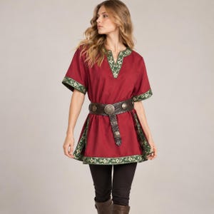 Op de afbeelding: Een donkerrode tuniek met korte mouwen en een V-hals, versierd met groen en wit bloemenborduursel langs de kraag en zoom. Het kledingstuk is in de taille ingesnoerd met een brede, donkerbruine leren riem met ronde metalen studs.