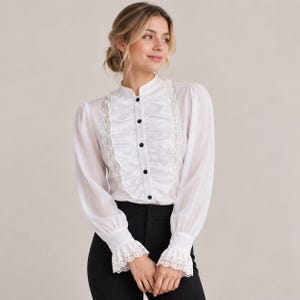 May include: A white long-sleeve blouse featuring a high collar, a central vertical ruffle panel, and delicate lace trim along the ruffles and cuffs. The garment includes a front button-down closure with small black buttons and long sleeves.