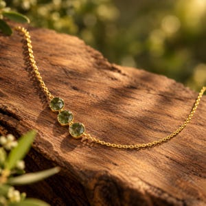 May include: A delicate gold chain necklace featuring three round, faceted olive green gemstones set in gold bezels. The necklace is arranged in a linear sequence, with the stones positioned centrally along the thin, polished gold metal link chain.