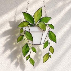 May include: A stained glass hanging plant featuring a white geometric pot and multiple trailing green leaves. The piece is suspended by a thin metal chain, with varying shades of translucent green glass creating the foliage and opaque white glass for the pot.