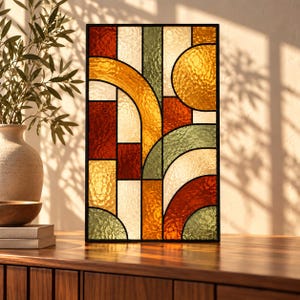 Puede incluir: Un panel rectangular de vidriera con una composición geométrica de piezas de vidrio texturizado en tonos ámbar, rojo intenso, verde oliva y crema. El diseño incluye arcos circulares y líneas rectas que se cruzan, enmarcado en metal negro.