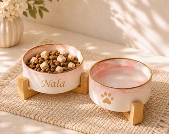 May include: Two round ceramic pet bowls with a pink and white marbled finish and gold rims, each resting on a light wooden stand. One bowl contains brown kibble and features the name Nala in gold, while the other is empty with a gold paw print.