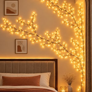 Puede incluir: Una lámpara de pared decorativa que consiste en tallos delgados, oscuros y ramificados adornados con numerosas luces LED pequeñas de tonos cálidos. Las ramas están dispuestas sobre la pared encima de una cama, creando una silueta brillante.