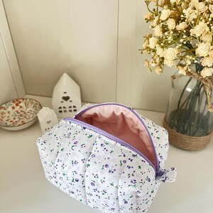 Puede incluir: Un estuche de tela acolchada con base blanca y un pequeño estampado floral morado. El estuche tiene un forro interior rosa claro y un cierre de cremallera morado. La estructura rectangular es suave y tiene costuras verticales.