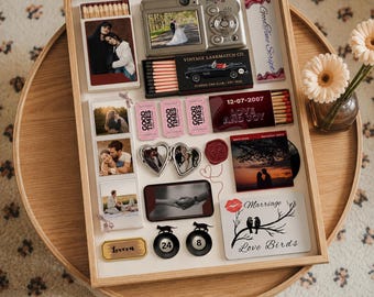 Könnte beinhalten: Ein hölzerner Schattenkastenrahmen mit einer Sammlung personalisierter Gegenstände, darunter eine Digitalkamera, Streichholzschachteln mit Fotos, ein "Good Time Scripts"-Feuerzeug, eine "Vintage Larkmatch Co."-Box, ein herzförmiges Medaillon, eine "Love and Joy"-Streichholzschachtel und ein "Marriage Love Birds"-Schild.