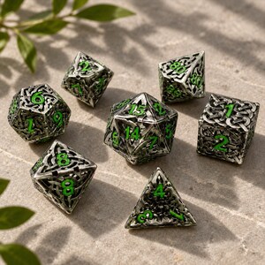 May include: A set of seven polyhedral dice featuring an intricate silver-toned metal filigree design with cut-out patterns. Each die has bright neon green numbers recessed into the metal structure, including a d20, d12, d10, d8, d6, d4, and a percentile die.
