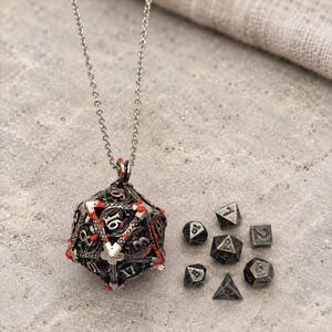 Può includere: Una collana in metallo color argento con un grande ciondolo a forma di dado a 20 facce, traforato e decorato con accenti in smalto rosso, accompagnato da un set di sei dadi poliedrici in metallo massiccio di varie forme, tra cui un cubo e una piramide.