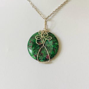 Può includere: Una collana color argento con un ciondolo rotondo in pietra verde e rossa. La pietra è avvolta in un design decorativo in filo metallico. La catena è anch'essa color argento, creando un gioiello semplice ma elegante.
