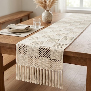 Peut inclure: Un chemin de table en crochet de couleur crème présentant un motif en damier alternant des carrés pleins et ajourés. Le chemin de table est terminé par de longues franges droites le long du bord inférieur, créant un aspect texturé.