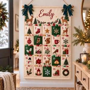 May include: A fabric wall-hanging advent calendar with the name Emily printed at the top. It features twenty-five numbered pockets decorated with embroidered holiday motifs like deer, trees, snowflakes, and gifts, accented by dark green velvet bows.