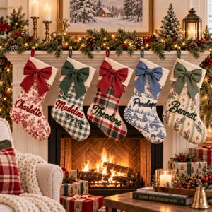 May include: Five personalized Christmas stockings hanging from a mantel, each featuring a large fabric bow at the top. The stockings display unique patterns including red floral, green gingham, red plaid, blue winter landscape, and striped pine branch designs.