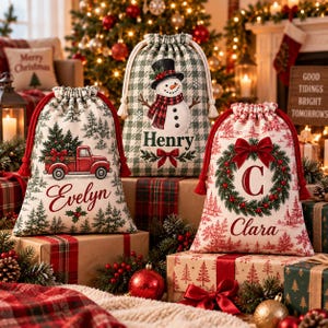 May include: Three fabric drawstring gift bags with holiday designs. One features a red truck with a tree, one a snowman with the name Henry, and one a wreath with the letter C and the name Clara. Each bag has a unique festive pattern.