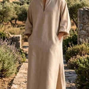 May include: A beige, ankle-length tunic made of a textured fabric, featuring long sleeves, a V-neckline, and side slits. The garment includes two side pockets and has a loose, straight-cut silhouette that falls to the lower calf area.