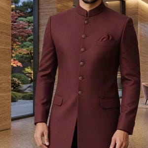 Puede incluir: Una chaqueta bandhgala larga de color burdeos intenso con cuello mao alto, tapeta frontal con botones circulares a juego, mangas largas y un detalle de pañuelo en el bolsillo del pecho, sobre pantalones oscuros.
