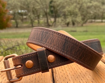 May include: A brown leather belt with a copper-colored buckle and rivets. The belt is made of distressed leather and has a stitched edge. The belt is lying on a wooden surface.