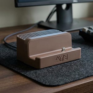 Peut inclure: Une station de charge marron mat pour console de jeu portable, dotée d'un connecteur USB-C central et du logo AYN gravé sur la base avant. L'unité présente un design rectangulaire avec un support arrière incliné et une section ventilée nervurée.