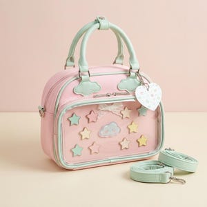 May include: A light pink handbag with mint green trim and handles. The front features a clear pocket containing star-shaped cutouts, a small white lace bow, and a central cloud graphic. A heart-shaped tag and two detachable mint green straps are included.