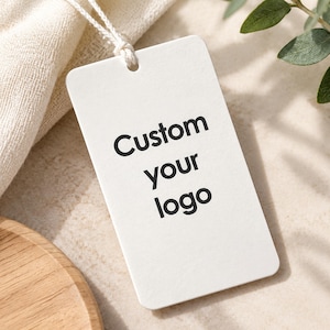 Op de afbeelding: Een rechthoekig wit papieren label met afgeronde hoeken, voorzien van de zwart gedrukte tekst Custom your logo. Het label is bevestigd aan een wit touwtje en heeft een strak ontwerp voor branding en identificatiedoeleinden.