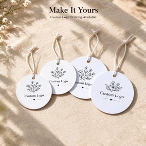 May include: Four white circular tags with string loops, each featuring a black line-art floral illustration, the text "Custom Logo," and a small heart symbol. The top of the image displays the text "Make It Yours" and "Custom Logo Printing Available."