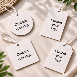 Op de afbeelding: Vier off-white papieren labels in ronde, rechthoekige, vierkante en afgeschuinde vormen. Elk label is voorzien van een wit touwtje en de zwarte tekst Custom your logo die gecentreerd op de voorzijde van het karton is gedrukt.