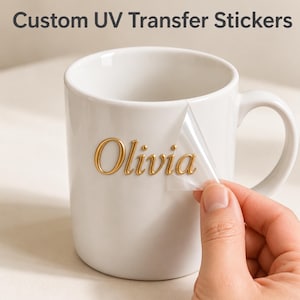 以下が含まれることがあります： 白いセラミック製のマグカップに、金色の筆記体でOliviaと書かれたカスタムUV転写ステッカーが貼られています。透明な粘着フィルムがマグカップの表面から部分的に剥がされており、その下の盛り上がった金色の文字が見えています。