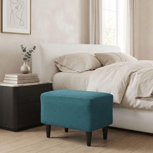 Op de afbeelding: Een rechthoekige voetenbank met een getextureerde blauwgroene stoffen hoes met een ruitpatroon, ondersteund door vier donkerbruine taps toelopende houten poten. De voetenbank staat voor een bed met lichtgekleurd beddengoed en een wit hoofdeinde.