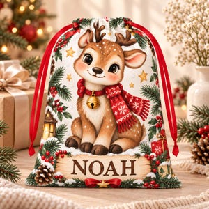 Op de afbeelding: Een witte cadeauzak met trekkoord met een bruin reekalfje met een rode sjaal en een gouden bel. De zak is versierd met dennentakken, rode bessen, sterren en een lantaarn. De naam NOAH staat in bruine letters op een banner onderaan.