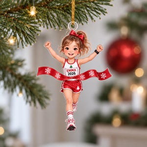 May include: A clear acrylic Christmas ornament featuring a cartoon runner with a ponytail and red bow, wearing a white tank top with a small green tree and the text EMMA 2026, red shorts with a white tree, and red sneakers, breaking a red ribbon.