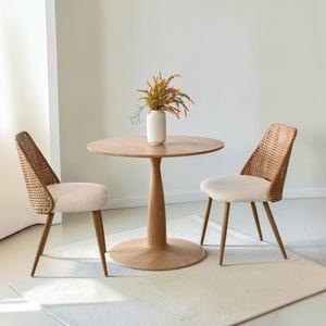 May include: A round wooden pedestal table with a light brown finish, accompanied by two chairs featuring woven rattan backs, cream-colored upholstered seats, and tapered wooden legs. A white ribbed vase holds orange flowers on the table.
