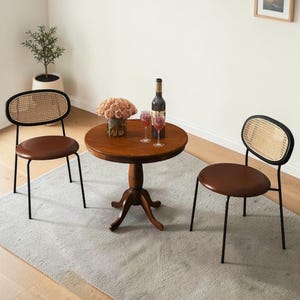 May include: A round wooden pedestal table with a dark brown finish, accompanied by two chairs featuring black metal frames, brown circular seats, and oval backrests with woven cane detailing. A wine bottle and two glasses sit on the table.