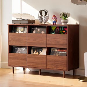 May include: A brown wooden storage cabinet with six open compartments for vinyl records, featuring a flat top surface holding a record player, headphones on a stand, a small figurine, and a vase with flowers, supported by four tapered legs.